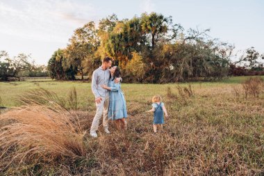 Young couple with cute two-years-old girl standing on the grass hugging each other in blue outfit