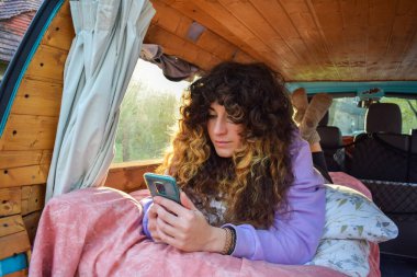Bağımsız kıvırcık saçlı bir kadın, yolculuk boyunca minibüsünün yatağında dinlenirken cep telefonuna mesaj atıyor.