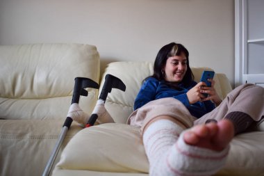 Young woman on couch at home with crutches and orthopedic plaster chating on the phone. Fracture of the broken leg foot or knee. Concept of rehabilitation and healing. Orthopedics and Traumatology.