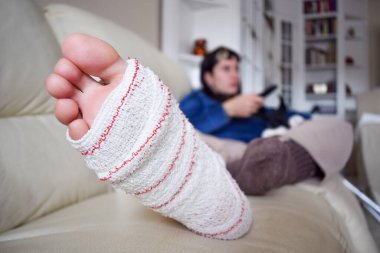 Young woman on couch at home with crutches and orthopedic plaster watching TV. Fracture of the broken leg foot or knee. Concept of rehabilitation and healing. Orthopedics and Traumatology.