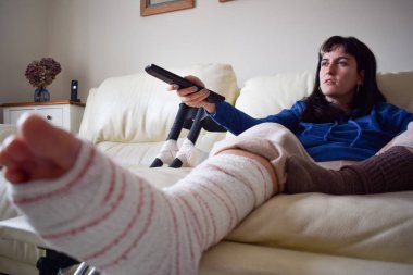 Young woman on couch at home with crutches and orthopedic plaster watching TV. Fracture of the broken leg foot or knee. Concept of rehabilitation and healing. Orthopedics and Traumatology.