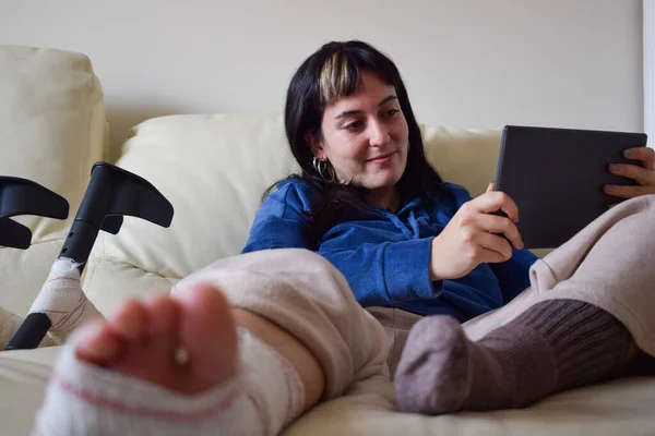 Young woman on couch at home with crutches and orthopedic taking selfie on tablet. Fracture of the broken leg foot or knee. Concept of rehabilitation and healing. Orthopedics and Traumatology.