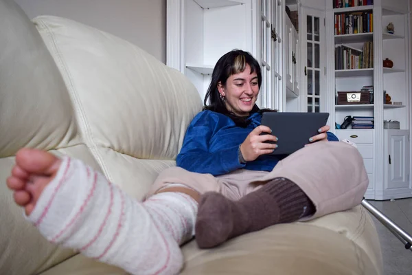 Young woman on couch at home with crutches and orthopedic watching series on tablet. Fracture of the broken leg foot or knee. Concept of rehabilitation and healing. Orthopedics and Traumatology.