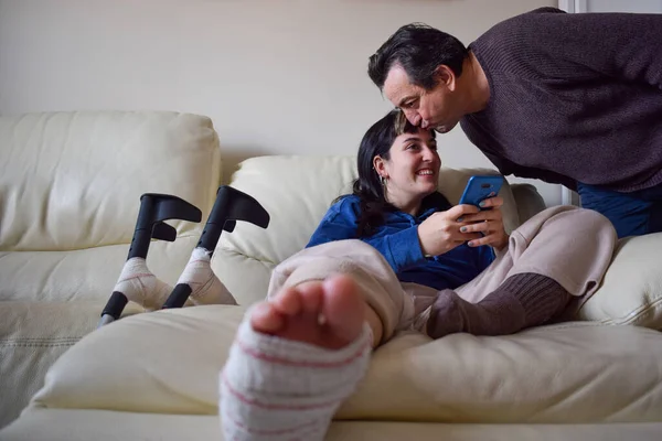 Father kissing her young woman daughter on couch with crutches in orthopedic plaster chatting on the phone. Broken leg foot or knee. Rehabilitation and healing concept. Orthopedics and Traumatology.