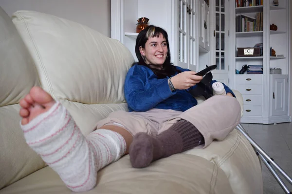 Young woman on couch at home with crutches and orthopedic plaster watching TV. Fracture of the broken leg foot or knee. Concept of rehabilitation and healing. Orthopedics and Traumatology.