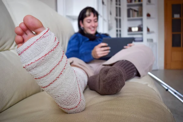 Young woman on couch at home with crutches and orthopedic watching series on tablet. Fracture of the broken leg foot or knee. Concept of rehabilitation and healing. Orthopedics and Traumatology.
