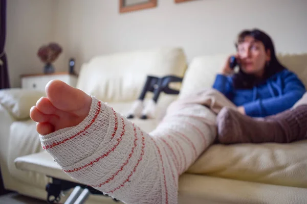 Young woman on couch at home with crutches and orthopedic plaster talking on the phone. Fracture of the broken leg foot or knee. Concept of rehabilitation and healing. Orthopedics and Traumatology.