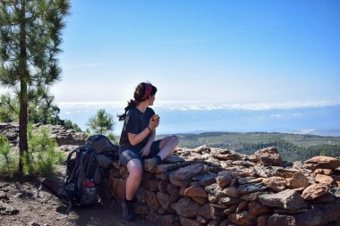 backpacker girl having Breakfast on edge of a top cliff with vale view. Traveller eating nourishing food. Trip along the ridge. Morning lifestyle. Apple for a tasty healthy snack meal in a hill hike