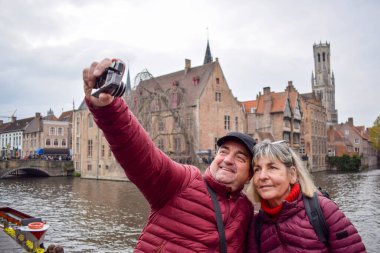 Yetişkin bir çift, Belçika, Bruges 'da bir kanalı olan ünlü bir turist bölgesinin arka planında selfie çekiyor. Avrupa 'da Kanal Köprüsü' ndeki Turist Kafkas ailesi. Mutlu olgun çift kıyafetleri.