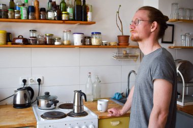 Uykulu bir adam, evde bir mutfakta dururken sabah kahvesinin hazır olmasını bekliyor. Kızıl saçlı, gözlüklü ve uzun saçlı bir erkek. Klasik mutfak..