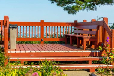 Red wooden and timber balcony with bench and hand rails with sky lined overlook midday in sun with foreground plants and tree. Steps and trash can in public area with deck for standing or smoking in suburban area. clipart