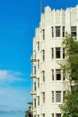 Ön bahçesinde yeşillik olan beyaz, uzun bir apartman ve gün ortasında bulutlu mavi gökyüzü arka planlı ağaçlar. Şehir merkezinde, kentte ve banliyöde beyaz bulutlar olan bir yerde..