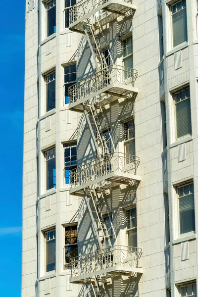 High rise beige apartment building with metal saftey fire escape ladder with windows and blue sky background in sun midday. Downtown in the neighborhood or in an urban setting with building in city.