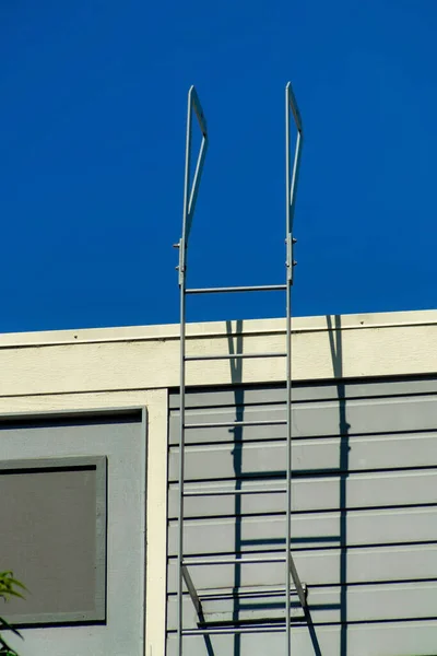 Emergency fire escape ladder on the side of modern building with blue and white wooden or timber trim. Midday in sun with bright blue sky background on flat roof in an urban or suburban area.