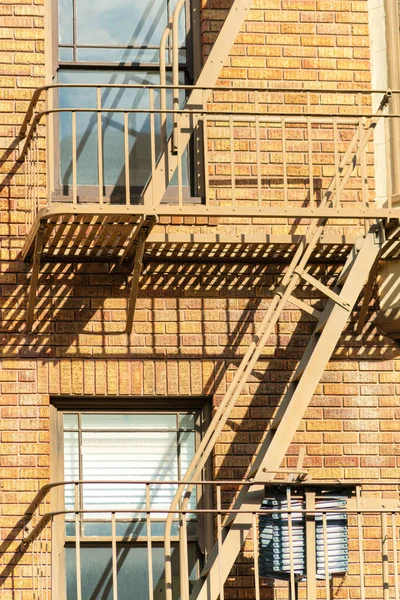 Beige or brown fire escape ladder on the side of brick building in a modern part of the downtown city. In midday sun with orange accent colors and windows on the exterior of the building.