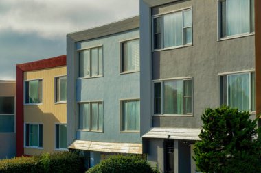 Akşamüstü güneşli, bulutlu ve gri gökyüzü arka planı mahallede ya da banliyöde olan kare şeklinde Coloful evleri. Ön bahçedeki ağaçlar ve evlerin yüzlerindeki gölgelerde çizgiler.