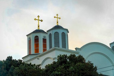 Çift katlı kilise kulesi, beyaz boya, kırmızı ve mavi iç döşemeler ve bir kilise binasının çatısında altın metal haç. Ön bahçe ağaçları ve gri ve sisli arka planlı bitkiler.