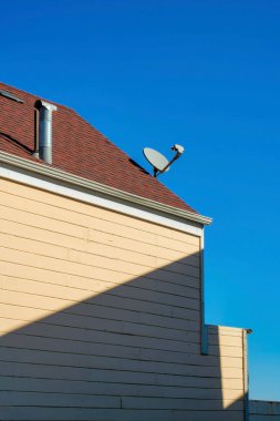 Dış yüzeyinde gölgelik çizgiler, ahşap sürahiler ve uydu ve baca delikleri olan kırmızı kiremitlerle ıssız bir ev cephesi. Öğleden sonra güneşinin altında açık mavi gökyüzü arkaplanı var..