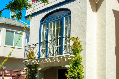 Küçük metal dekoratif balkon, beyaz bina cephesi, sarmaşıklar, ön bahçe ağaçları ve yeşillikler. Öğleden sonra güneşinde mavi aksanlı cam kapı ve mavi gökyüzü boyalı.