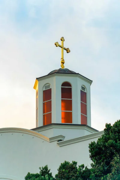 Church steeple with red interior and gold metal cross on roof with white exterior paint on chruch building in late afternoon clouds and sky. In the church courtyard with trees in foreground.