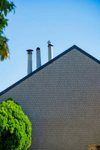 White house facade with wooden tiles and visible chimney pipes in late afternoon shade in the city. In a suburban setting in the neighborhood with back yard trees and bue sky background.
