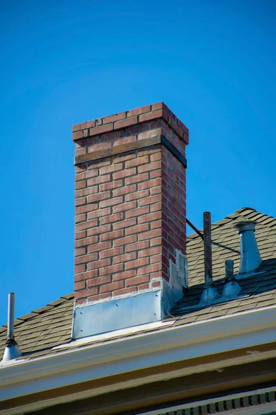 Red brick chimney on brown roof with decorative tiles and metal chimney pipes on double gable blue sky house or home. In the neighborhood or in the city with midday sun in the metropolis.
