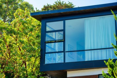 Geniş panoramik pencereli modern bir ev ve düz çatılı bir kutu ve öğleden sonra güneşinde kutu şeklinde. Ön bahçedeki ağaçlar ve mavi gökyüzü arka planı şehirde ya da mahallede..