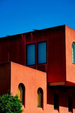 Blok tarzı inşaatın yakın görüntüsü ya da kırmızı kahverengi sıva dış görünüşlü mimari ya da görünür pencereli ve ön bahçe ağaçlı cephe. Öğleden sonra güneşinde ve açık mavi arkaplanlı gölgesinde.