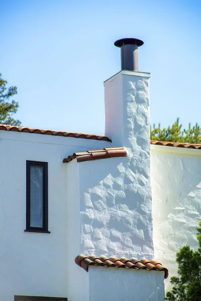 Tall chimney with white stucco exterior and adobe red roof tiles with back yard trees and clear blue or white gradient sky. Late in the afternoon in neighborhood or in the suburban part of the city.