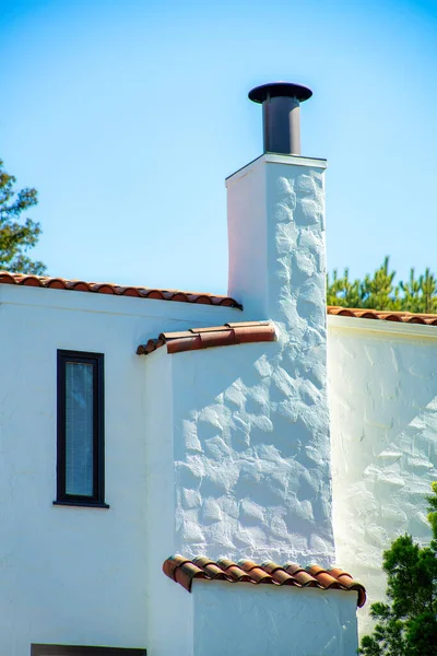 Tall chimney with white stucco exterior and adobe red roof tiles with back yard trees and clear blue or white gradient sky. Late in the afternoon in neighborhood or in the suburban part of the city.