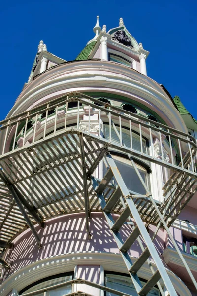 stock image Gray metal fire escape ladder and balcony with white decorative building facade and clear blue sky background in sun. Late in the day in the urban part of the downtown city San Francisco.