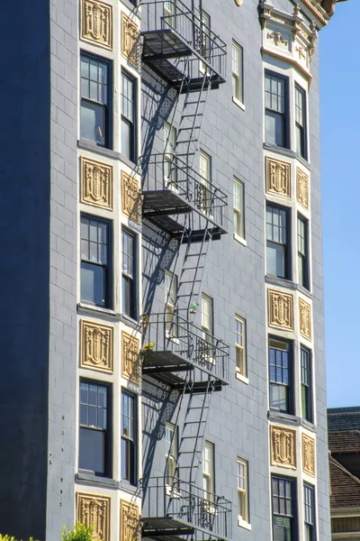 Blue building facade with yellow decorative exterior and black fire escape ladder and balcony in late afternoon sun with clear blue sky. In the urban part of city on building face with stucco walls.