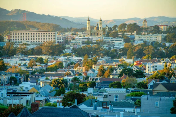 Kaliforniya 'nın merkezindeki San Francisco semtlerinde gün batımında dağlarla birlikte altın ve turuncu şehir manzarası yayılıyor. Şehir meydanlarında ve iş merkezinde görünür ağaçlar ve eğimli gökyüzü.