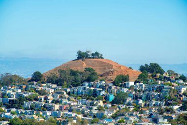 San Francisco California 'nın şehir merkezindeki tepedeki kırsal bölgede ağaçlar, dağlı kuru otlar ve gün içinde sisli gökyüzü. Mahallede evleri olan mavi ve beyaz eğimli arka plan.
