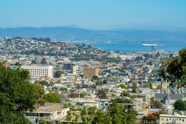 San Francisco şehir merkezinde sisli dağları, mavi gökyüzü, okyanus arkaplanı ve şehir ağaçlarıyla yayılmış. Öğleden sonra güneşinin altında evler ve evler var..