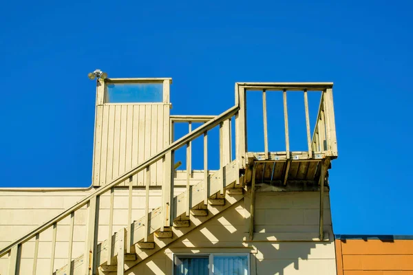 Stairs leading to the roof of the house to a flat patio with beige or yellow paint with blue dark sky in the late afternoon sun and shade. In a suburban area of the neighborhood roof access on top.
