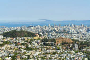 San Francisco şehir merkezinde tepeler, ağaçlar ve güneş altında ağaçlar ve ağaçlar. Evleri ve komşuları var. Arka planda okyanus ve mavi ve beyaz gökyüzü var.