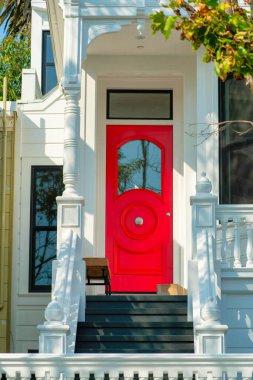 Red painted front door with glass window and grey steps with white traditional hand railing and picturesque front porch with hanging plant in specled midday sun. Suburban neighborhood afternoon. clipart