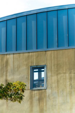 Mavi metal rendeleyici aksanlı yuvarlak çatı ve gölgede küçük bir penceresi olan pürüzsüz beton duvarlar. Öğleden sonra ilerleyen saatlerde gökyüzü mavi ve ön planda yeşilliklerle kasıla kasıla yürürken.