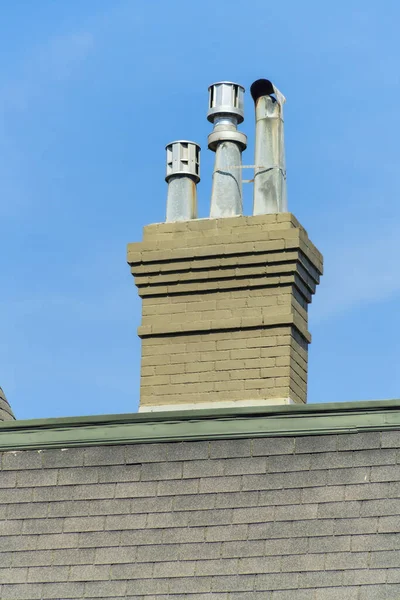 Modern chimney with stucco brown exterior on gray slatted roof in midday sun with metal chimney pipes. Blue sky with light clouds in the neighborhood of the suburbs or city.