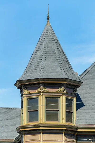Exterior turret or spire with beige stucco home or house and gray slatted roof with spike on pinacle. Blue sky background with some white clouds in late afternoon sun in the neighborhood or suburbs.