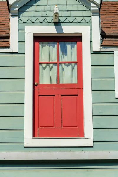 Red hidden door on side of building with white trim on the frame and green gray exterior horizontally slatted wood or timber house or home. In midday sun with brown roof in the city or neighborhood.
