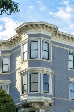 Mavi ahşap ev cephesi, yatay ahşap sürahiler ve mavi gökyüzü arka planlı ön bahçe yaprakları. Gün ortası gölgesinde, bulutlar, beyaz aksanlı boya ve binaların çatısında.