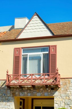 Kahverengi çatılı, beyaz ve bej boyalı, kırmızı tüylü ve mavi gökyüzü olan küçük balkonlu tuhaf bir ev cephesi. Öğleden sonra güneş doğarken mahallede ya da şehirde binalarda..