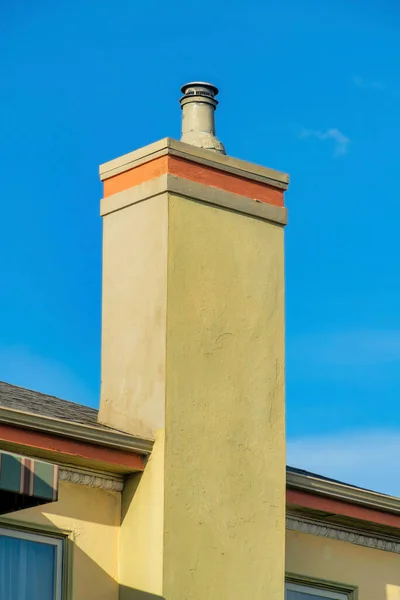 Cement chimney with beige stucco exterior with brick accent and metal pipes in late afternoon sun with blue sky background. Detail shot of house in the neighborhood or in suburban area.