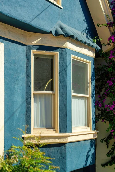 Bay window on blue stucco house with small tile roof and white accent paint around windows and glass on facade of home. In the neighborhood or in a suburban part of the downtown city in midday sun.