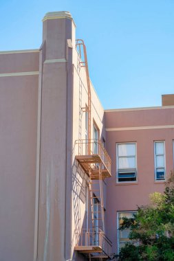 Yangın merdivenli pembe ya da turuncu sıva bina ya da mavi arka planda gölgesi olan güneşte balkondan kaçmak. Şehirde ya da arka bahçesi olan mahallede..