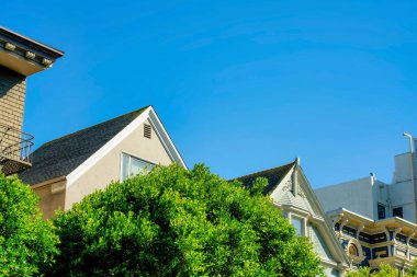 Gün ortası güneşinde ve ön bahçe ağaç tepe örtüsünde gable tarzı çatıları olan dekoratif sıva cepheli görünür çatılar. Öğleden sonra geç saatlerde. Mahallede temiz mavi fotokopi alanı var..