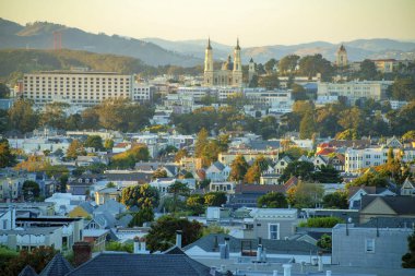 Kaliforniya 'nın merkezindeki San Francisco semtlerinde gün batımında dağlarla birlikte altın ve turuncu şehir manzarası yayılıyor. Şehir meydanlarında ve iş merkezinde görünür ağaçlar ve eğimli gökyüzü.