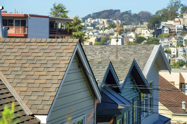 Rows of lit up roofs with brown tiles and double gable edges with deep suburban neighborhood background with front yard trees. In the back with chimneys and city area in san francisco california.